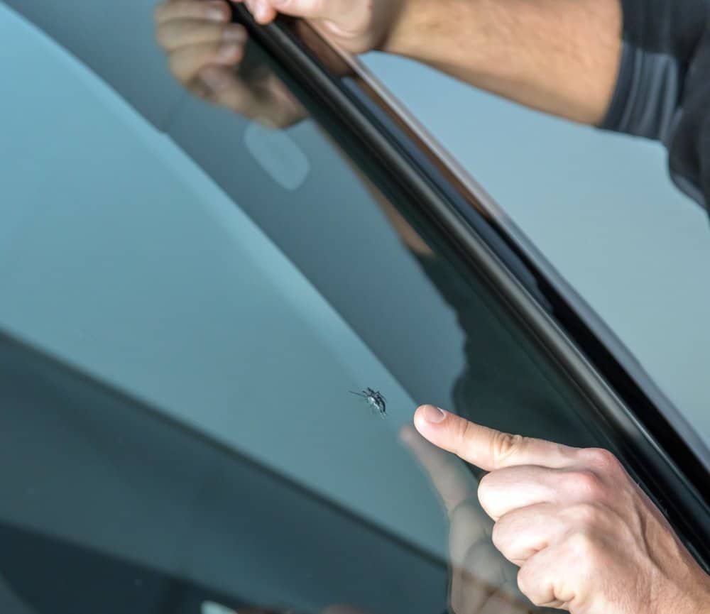 A person points to a small chip on a car windshield, clearly concerned. The glass reflects light, highlighting the damage. Their arm and hand suggest they might be considering windshield repair options to avoid more costly windshield replacement down the road.