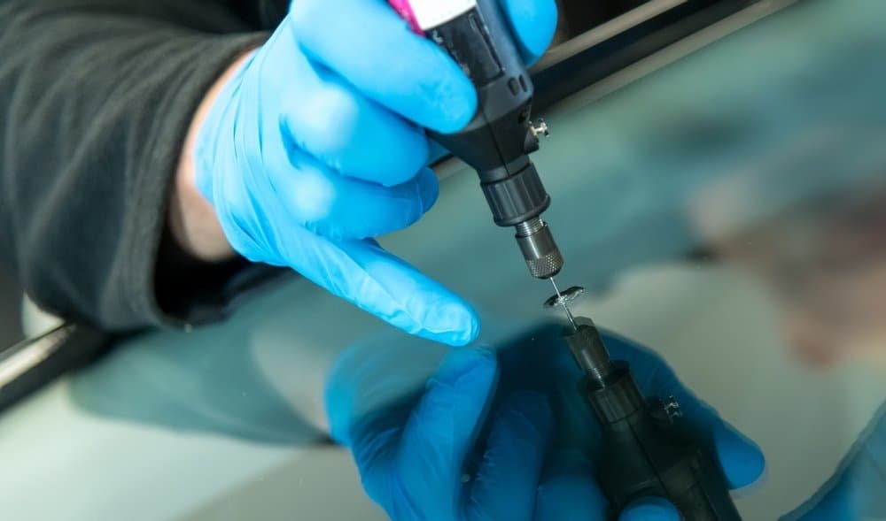 A person wearing blue gloves uses a drill tool to repair a crack in a car windshield while ensuring precise ADAS calibration.