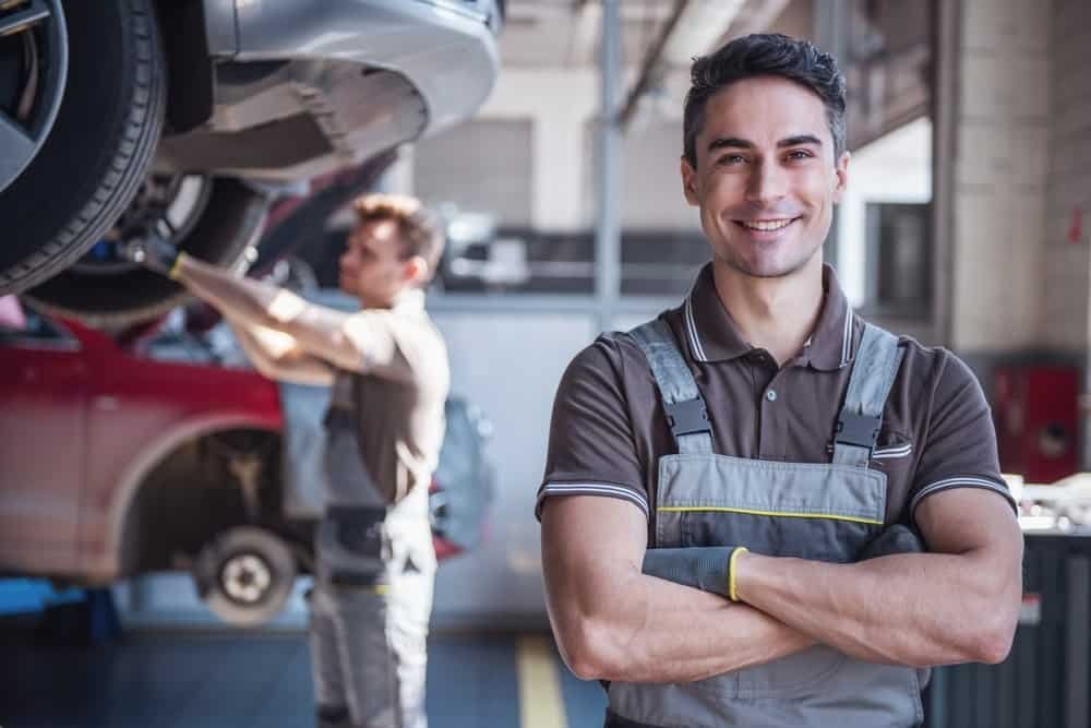 Two mechanics in a garage; one is smiling and standing with folded arms in the foreground, while another works on windshield repair for a lifted car in the background. Both are wearing grey overalls and brown shirts.