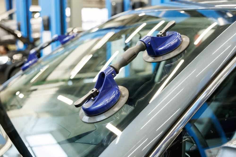 A close-up of a car windshield with two blue suction cups suggests a repair or installation in progress, possibly involving ADAS calibration. The background features a blurred view of garage equipment and blue supports.