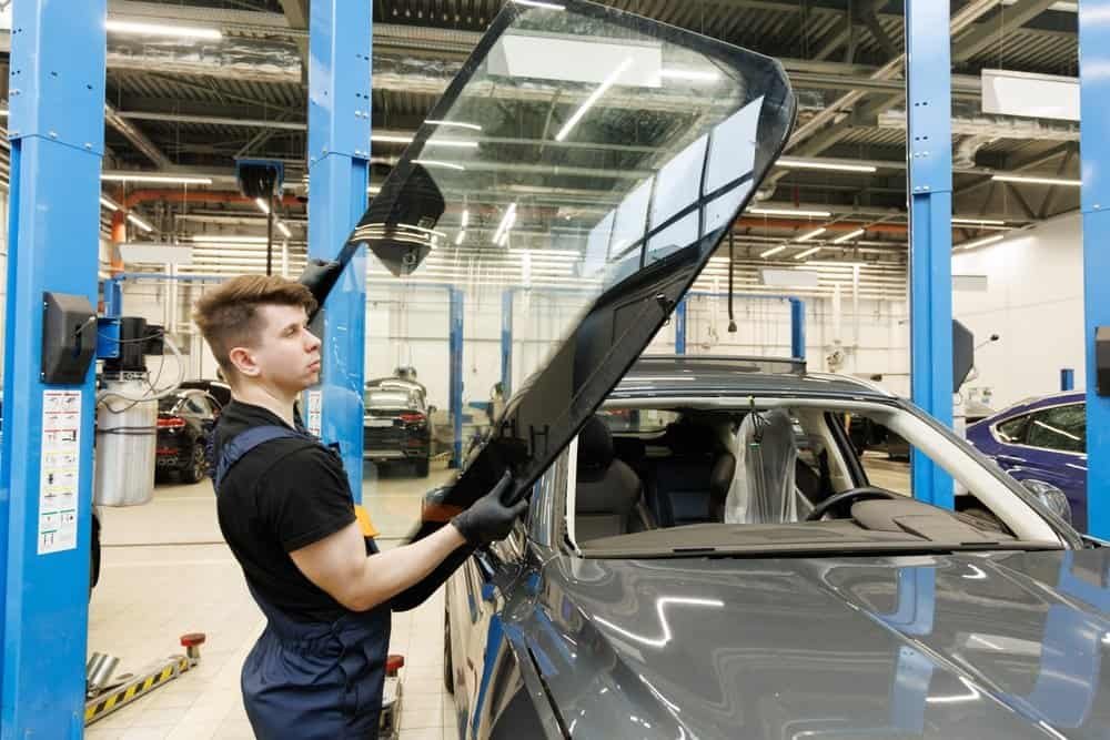 A garage worker expertly lifts a large car windshield, preparing for a seamless Windshield Replacement. Clad in a black shirt, blue overalls, and gray gloves, he stands amidst other cars and automotive equipment in the well-lit workshop.