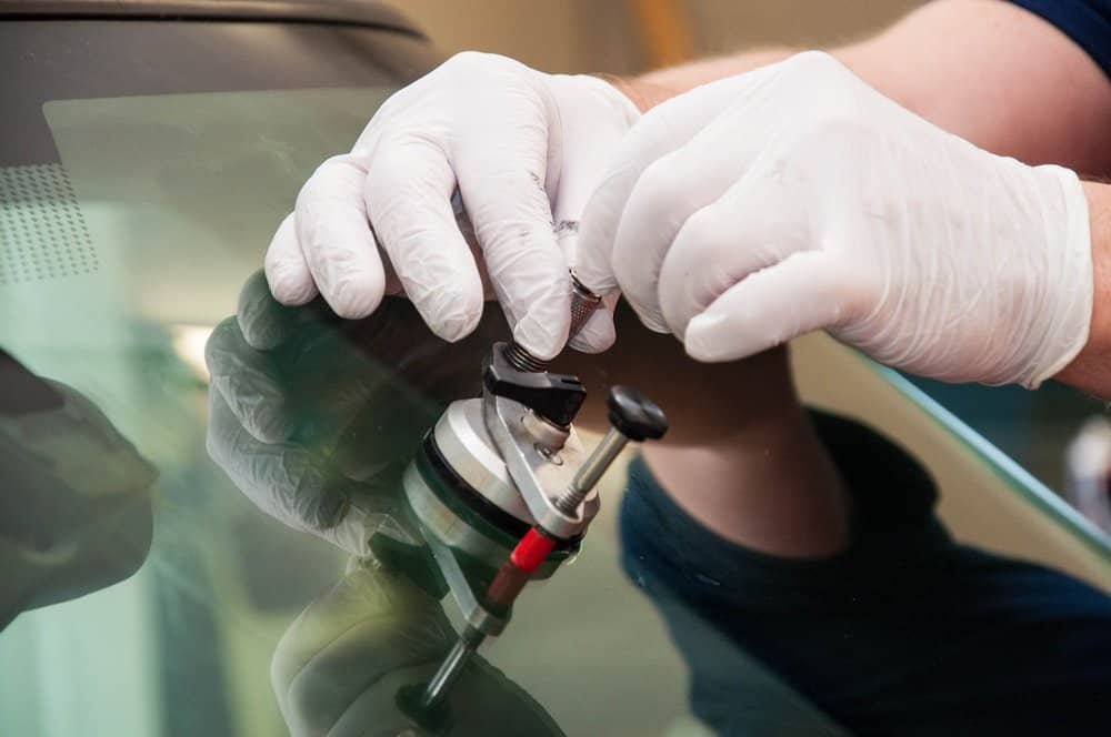 A person wearing white gloves meticulously repairs a car windshield using a specialized tool. The close-up image captures skilled hands focusing on rock chip repair, sealing a crack in the glass with precision and care.