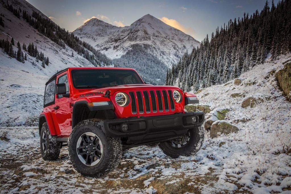 A red Jeep is parked on a snowy mountain trail surrounded by pine trees and snow-covered peaks.