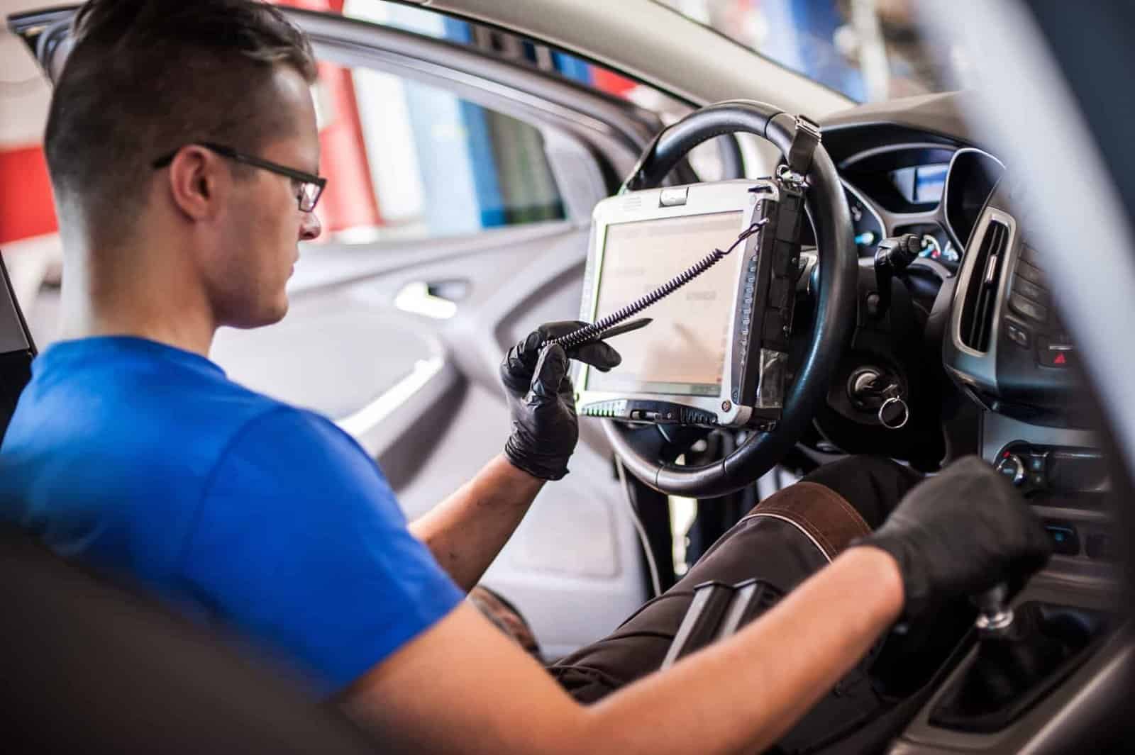 A person in a blue shirt and black gloves is seated in a car, using a diagnostic tablet attached to the steering wheel. The open car door allows a glimpse of the interior as they prepare for windshield repair.