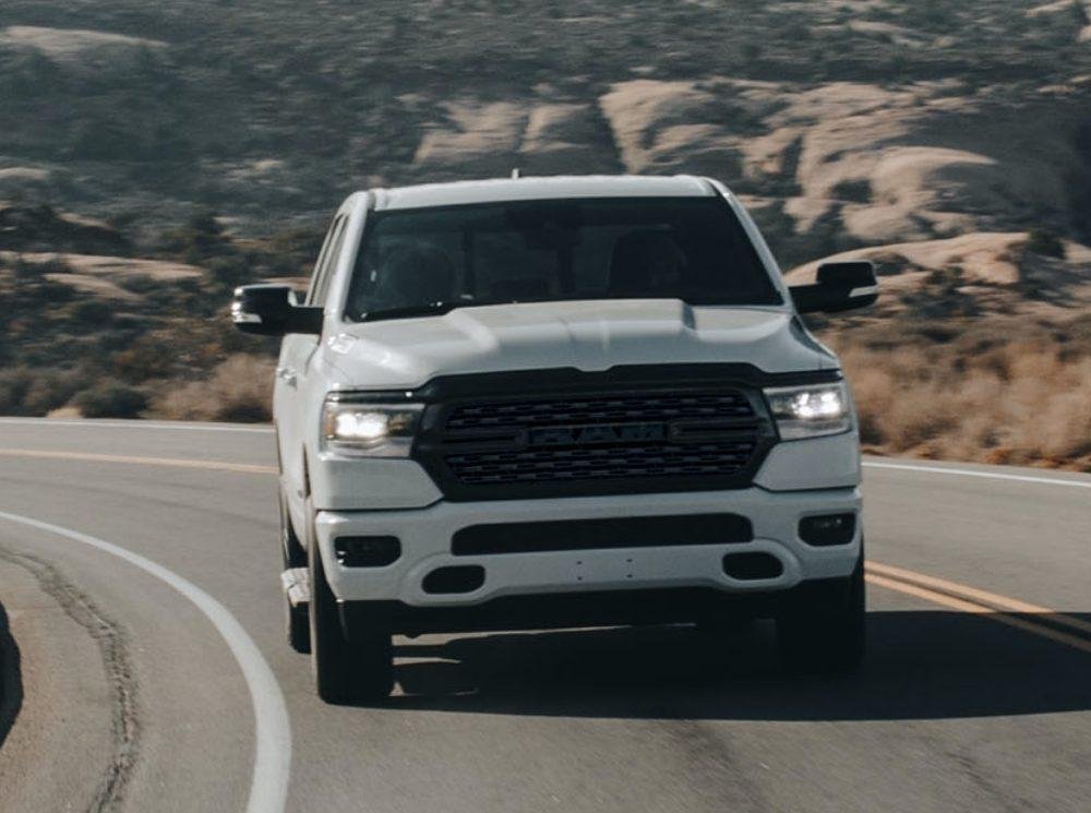 A white pickup truck drives on a winding road through rocky desert terrain under bright daylight.